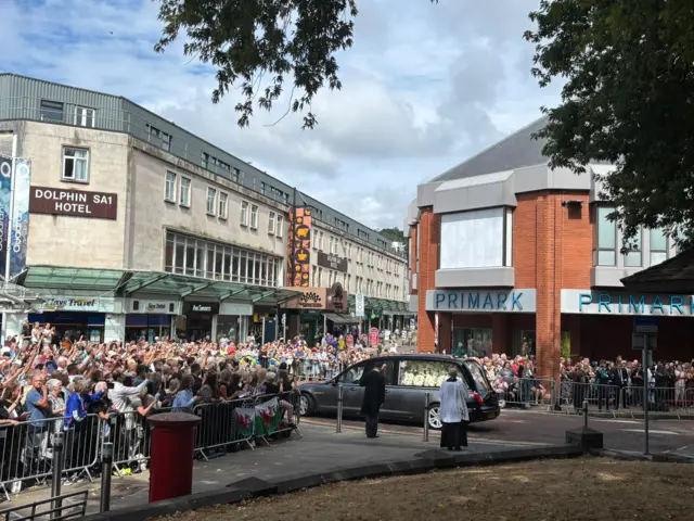 A funeral car with a coffin inside being driven through Swansea, surrounded by crowds