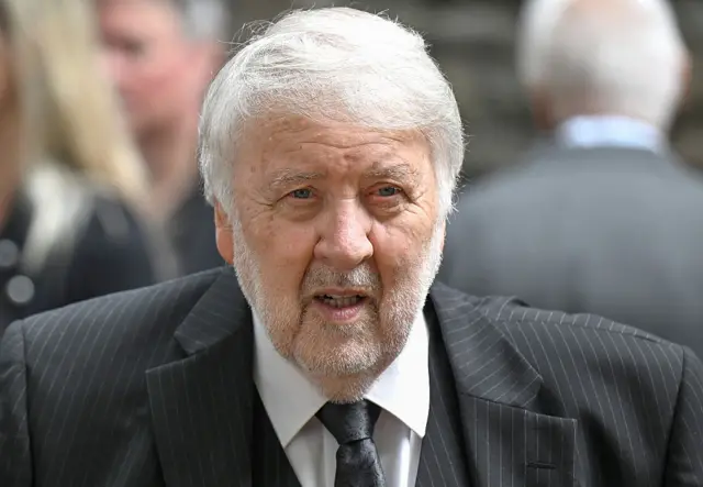 A man with white hair and a white stubble with a black and white pinstriped suit. It is a head and shoulders shot.