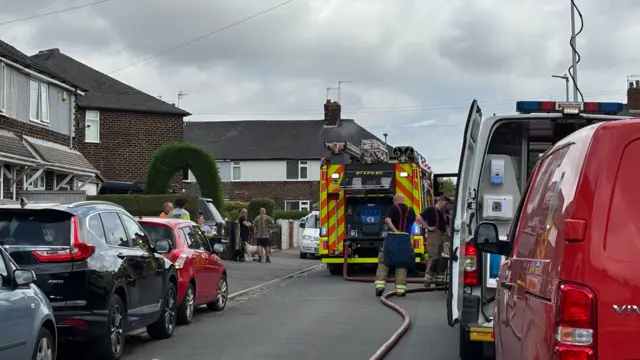 Firefighters and a fire engine at the scene of a fire on a residential street. Two crew members are at the back of the truck and a hose can be seen on the ground. Cars are parked either side of the road and people are on the left watching.