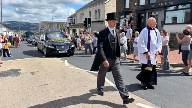 A man wearing a top and tail suit is walking in front of a hearse, alongside a reverend canon, on a street