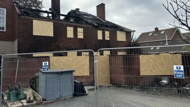 A boarded up building with a roof damaged by fire and metal fencing in front
