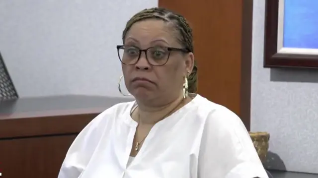 A black woman with glasses and a white shirt sitting in the witness stand in court.