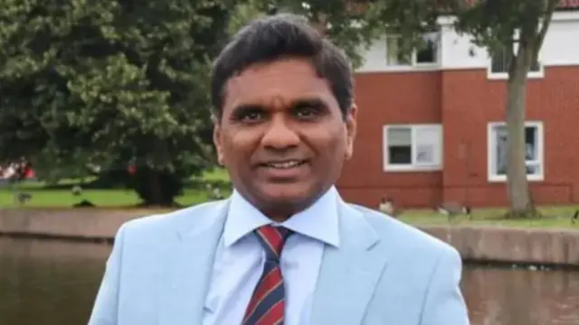 A man with dark hair and a pale suit with a red and blue striped tie in front of a canal