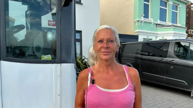 A woman with blonde hair wearing a pink strapped top. She is stood next to a black van.