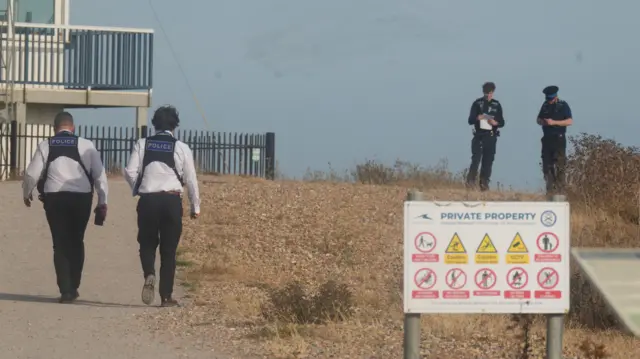 Police officers walk along a gravel path near the seafront while two other officers stand nearby during the emergency response close to Shoreham Port.