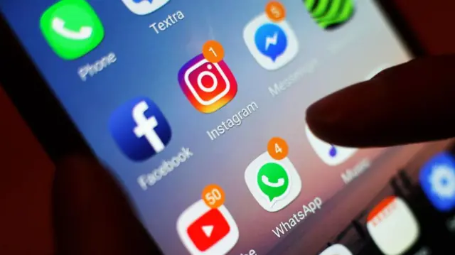 A close-up of a thumb on a smartphone screen with the Facebook, Instagram, YouTube and WhatsApp apps visible.