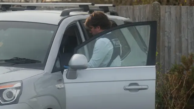 A police officer in a white shirt and police vest stands beside an open silver vehicle door while looking inside the vehicle. A wooden fence and vegetation can be seen in the background.