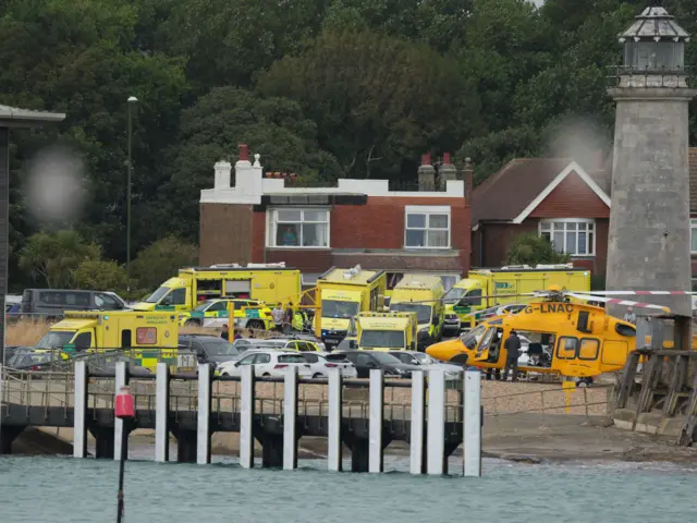 A number of ambulance vehicles parked by water. A helicopter is hovering above the water.