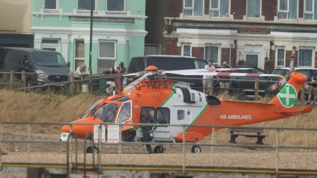 An orange and white air ambulance helicopter is parked on a beach area near Shoreham Port while emergency personnel stand nearby.