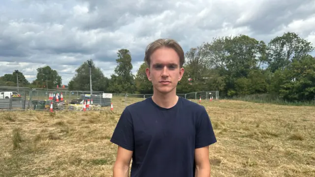 A student wearing a dark blue top stood in a field with a metal fence in it.