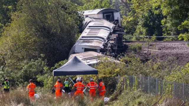 Railway carriages on their side, stretching from a railway line down the side of a small embankment. Emergency workers crowd at the bottom of the embankment.