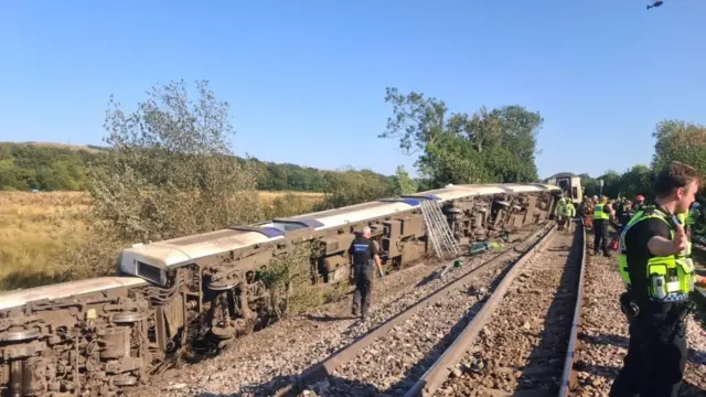 Three carriages of a train that have come off the track in Lewes and are lying on their side, while police officers work beside it