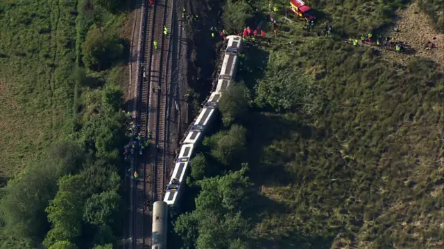 A train on its side by tracks with lots of people in hi vis walking around
