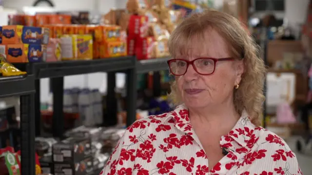 Penny is pictured in a flowery shirt in a foodbank with supplies behind her