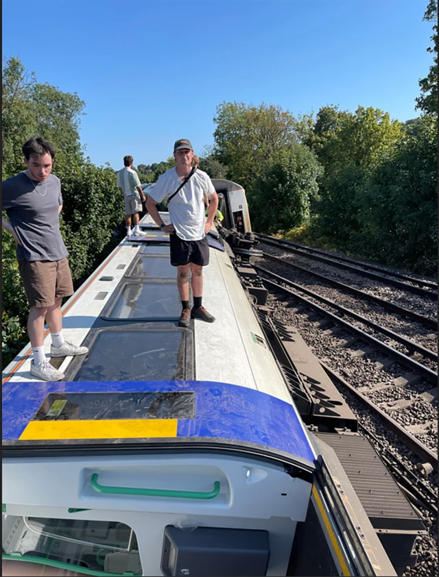 Three commuters of a train that tumbled off its tracks are standing on top of the one of the carriages which is lying on its side