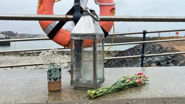 Flowers and a candle holder left next to a ring floatation device near Shoreham fort on the seafront