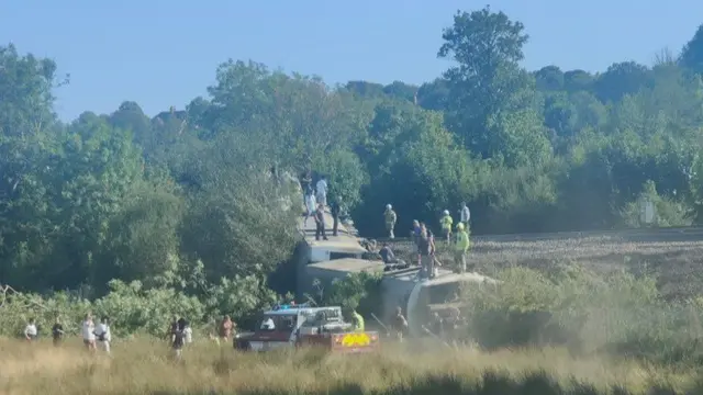 Image showing two overturned train carriages with several people standing on top of them, as well as emergency services personnel around them
