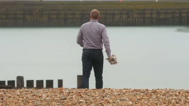 A man lays flowers on the beach in Shoreham-by-Sea, Sussex. He has his back to camera and is wearing black jeans and a purple shirt.