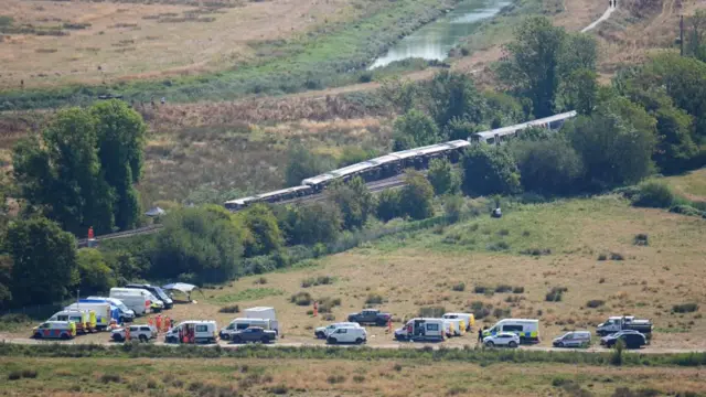Train tracks overturned on their side having fallen off the tracks while over a dozen emergency vehicles are parked nearby