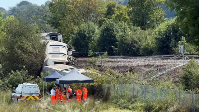 A train on its side next to an embankment with workers under a tent watching on