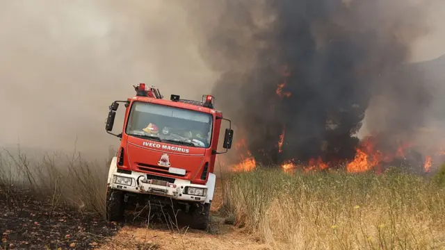A fire truck driving through a field with a huge red and black smokey fire blazing behind and throwing smoke into the air