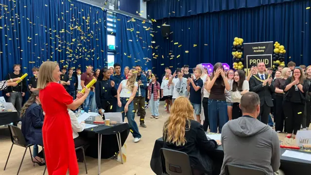 Confetti falls over a crowded school hall. People stand and applaud around tables, while a person in a red outfit operates a confetti cannon. A banner reading “Proud” and black-and-gold balloon displays are visible at the front.