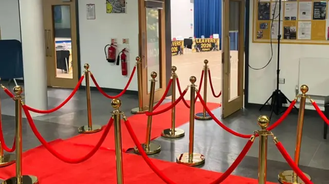 Red carpet entrance lined with gold stanchions and red ropes inside a school hall. Open double doors lead to an event space, with a grand piano, noticeboard and pop-art wall decorations visible in the foyer.