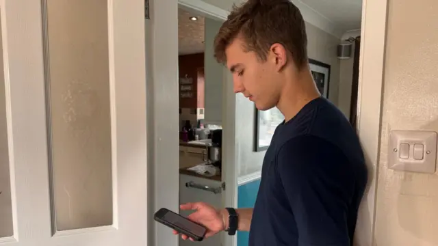 Person standing in a doorway inside a home, looking down at a smartphone. The person wears a dark blue T-shirt and a smartwatch. An open door reveals part of a kitchen in the background, with neutral-coloured walls and natural indoor lighting.
