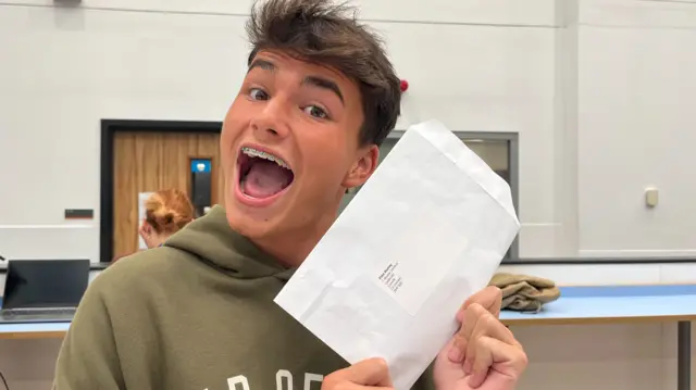 Finn holding up his exam results envelope with a big smile, wearing a green hoody with short brown hair