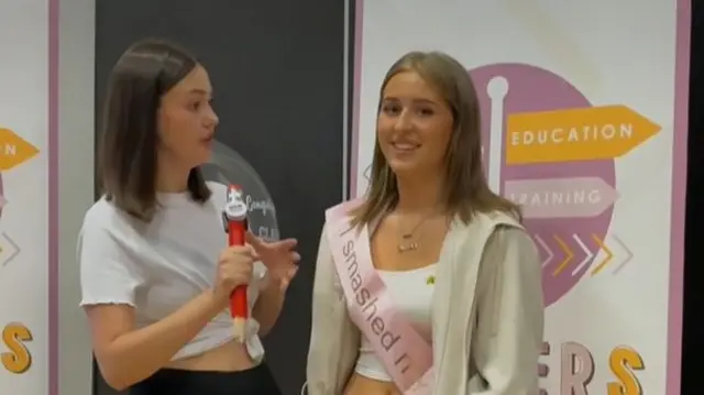 A woman wearing a white top and black skirt holds a microphone near a teenage girl who is wearing a beige hoodie and trousers with a pink sash that says 'I smashed my GCSEs I'm going to Ibiza'