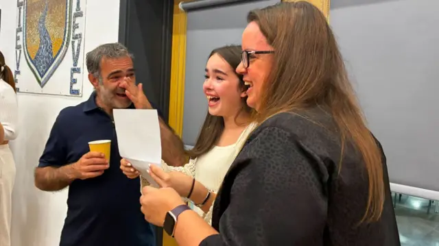 Several people stand near a doorway inside a school building, holding sheets of paper. A wall mosaic featuring the Leeds Brook Academy crest is visible behind them. One person holds a cup while the group gathers in conversation.