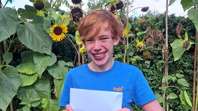 Charlie smiles and holds up a white envelope. There are several sunflowers behind him