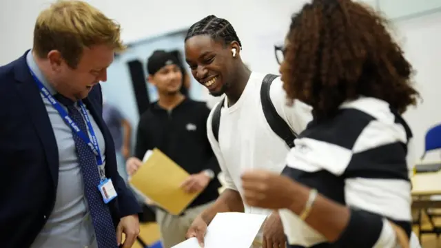 A teenage boy and girl open results in front of a teacher in a suit.