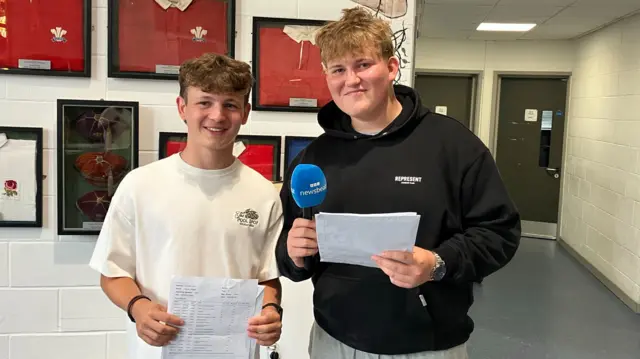 Jake and Haydn smile as they hold up their results in a school corridor. One of the boys is holding a blue microphone that says 'BBC Newsbeat'