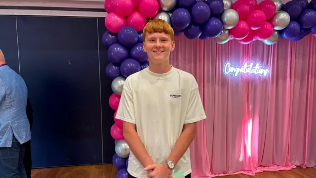 Reagan is seen holding a programme beneath a pink, purple and silver balloon arch at a celebration event. A neon “Congratulations” sign glows against pink drapes in the background, with a polished wooden floor and guests visible at the edge of the room.