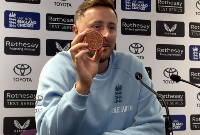 Ollie Robinson holds up half of the match ball in a press conference