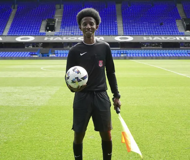 Nasser Hamid stands on a football field holding a flag and a football. He wears a black refereeing uniform.