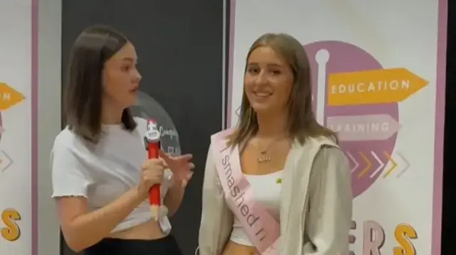 Isla in a t-shirt, zip up hoodie and sash speaking to a reporter.