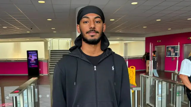 Bikram is pictured wearing a black hooded tracksuit standing between ticket barriers in a brightly lit indoor concourse. Pink walls, stairways and electronic displays are visible in the background, while transport staff stand near the barriers on the right.