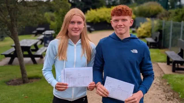 Dara and Kate standing outside holding their results slips, both with red hair and blue hoodies and in a grassy garden