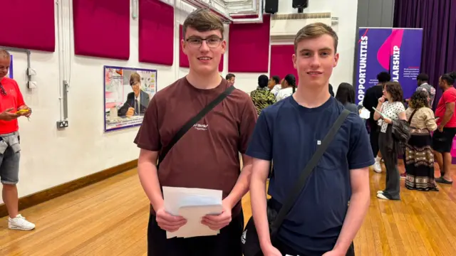 Twins Daniel and Joshua stand in a hall holding papers, with exhibition displays and visitors in the background. Bright pink acoustic panels line the walls, and banners reading “Opportunities” and “Scholarship” are visible, suggesting an education or careers event.