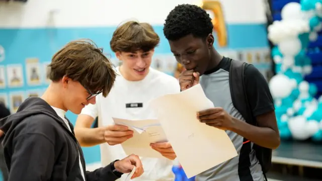 Three boys holding GCSE results in envelopes, looking at them and smiling or holding their hand to their mouth.