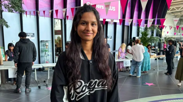 Amandeep is seen standing in the atrium of Sandwell College, facing the camera. Pink and white bunting hangs overhead, while tables and visitors are visible in the background. The person wears a black varsity-style jacket with white detailing.