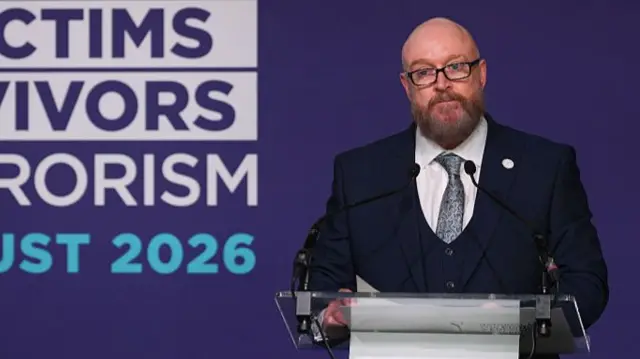 Paul Price is pictured speaking from a podium against a purple background.