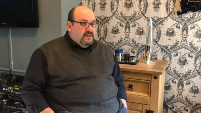 Paul Biddle is pictured speaking indoors, wearing a long-sleeved black shirt and grey vest.He has his hands in his pockets.