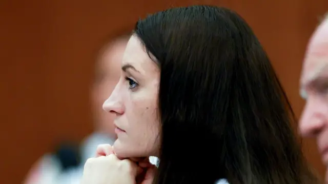 A woman sits with her head in her hands, looking straight ahead. We see her profile from the side. She has long dark hair, which frames a pale face and a chin that juts out slightly.