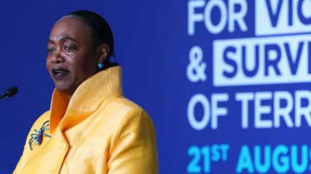 Thelma Stober is pictured speaking against a purple background, she is wearing a bright yellow jacket with a large spider brooch.