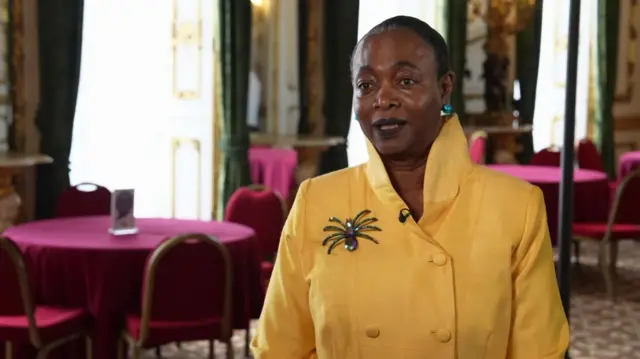 A black woman with short hair stares at someone off screen during an interview. She is wearing a yellow top with buttons running down the middle and a high collar.