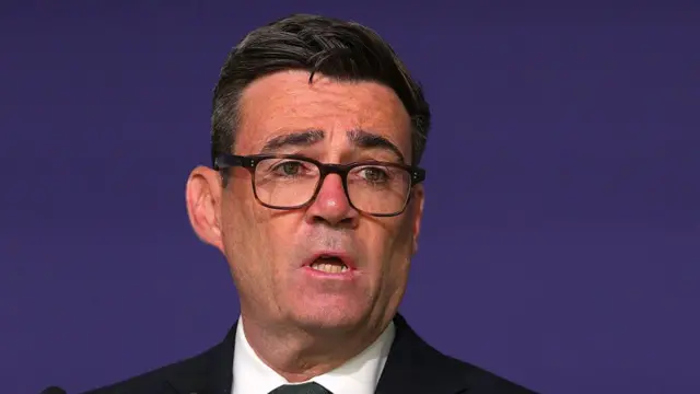 A close-up of Andy Burnham speaking against a purple background, wearing a suit and tie.