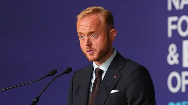 Travis Frain is pictured into microphones against a purple background.